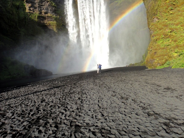 skogafoss waterfall black pebbly 2 tiny people in front of bright water falling arc of rainbow between water and rockface upper right